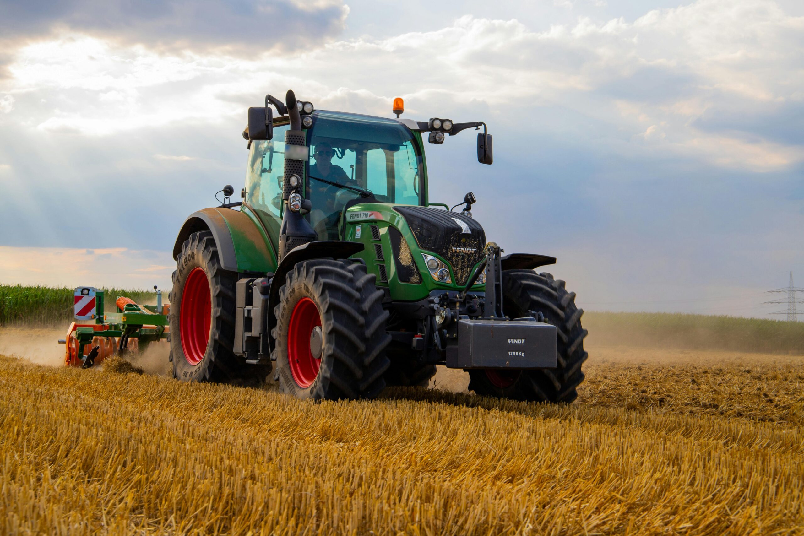 Home A powerful green tractor plowing a dusty wheat field under a cloudy summer sky in rural Germany.
